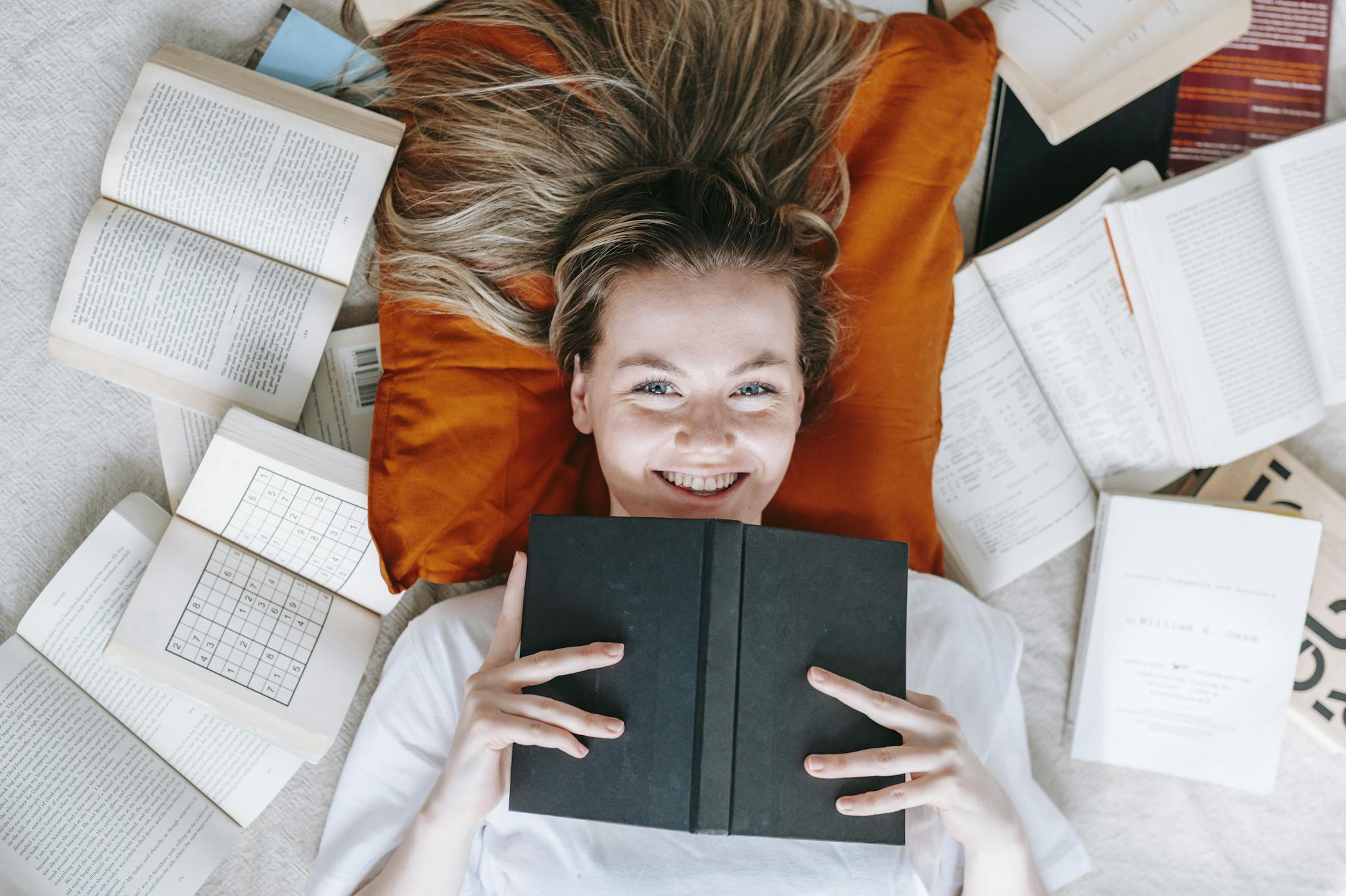 Top view of young female lying on floor among opened textbooks and books and smiling at camera